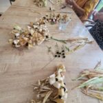 Na leseni mizi so razporejeni šopi rož, narejenih iz koruznih listov in črnih cvetnih sredic. Ob njih sedijo starejše osebe, ki izdelujejo dodatne cvetove. Na mizi so tudi kozarci z vodo in škarje.