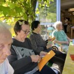 Starejši stanovalci doma sedijo pod pergolo, okrašeno z vinsko trto, in iz oranžnega papirja izrezujejo jesenske oblike.