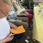 Stanovalec doma za starejše sedi za mizo in s škarjami izrezuje oranžen papir, ob njem sedijo drugi stanovalci.