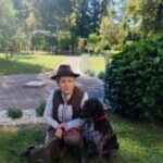 Ženska v lovski opravi s klobukom sedi na travi v parku poleg črno-belega psa na povodcu. V ozadju so drevesa, potka in zelenica.