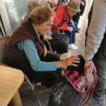Skupina starejših oseb sedi na klopih ob domu. Ena gospa sega z roko proti psu z rdečo rutko okoli vratu, medtem ko drugi nasmejano opazujejo.