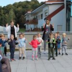 Otroci iz vrtca s spremljevalkama izvajajo gibanje z dvignjenimi rokami, za njimi sedijo starejši stanovalci.