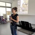 Ženska z očali in spetimi lasmi stoji ob električnem klavirju, drži knjigo in pripoveduje ob projekciji ilustracije na platnu.
