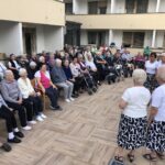 Skupina stanovalcev sedi zunaj na dvorišču doma starejših občanov in spremlja nastop pevskega zbora žensk v belih majicah in črno-belih krilih.