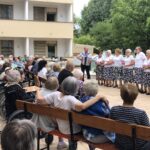 Pevski zbor žensk ob spremljavi harmonike nastopa pred stanovalci, ki sedijo zunaj na klopcah in invalidskih vozičkih, v ozadju so stavba in drevesa.