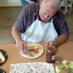 Stanovalec za mizo si na palačinko namaže marmelado, ob njem krožnik z zvitimi palačinkami.
