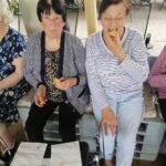 Dve stanovalke sedita druga ob drugi; ena gleda naravnost v kamero, druga z zaprtimi očmi ugrizne v kos kruha.