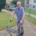 Starejši gospod v modri karirani srajci hodi z hoduljo po pločniku na vrtu doma.