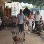 Stanovalci in otroci spremljajo igro z metanjem žoge v koš na dvorišču doma.
