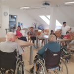 Skupina stanovalcev in otrok sedi za mizami v notranjem prostoru in se skupaj druži.