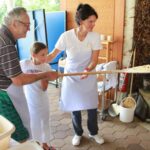 Starejši moški, deklica in ženska skupaj polagajo pico na leseno lopato pred krušno pečjo.