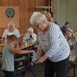 Starejša gospa in deklica skupaj sodelujeta pri igri s skodelico, okoli njiju sedijo stanovalci doma in otroci.