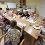 Skupina otrok sedi okoli velike mize in skupaj je zajtrk iz skodelic in kruha. Na mizi so tudi napitki in škatle z živili.