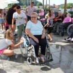 Nasmejana gospa v invalidskem vozičku, ki ji otroci in mladi pomagajo pri igri z vodo, medtem ko jo drugi udeleženci opazujejo.