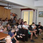 Otroci sedijo v notranjem prostoru in jedo sladoled, medtem ko skupaj gledajo projekcijo na platnu.