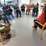 Skupina stanovalcev sedi v krogu in posluša predavateljico, ki drži mikrofon.