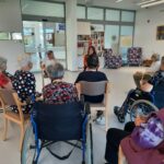 Stanovalci v invalidskih vozičkih in na stolih poslušajo predavanje v večnamenskem prostoru doma, predavateljica sedi spredaj in drži mikrofon.