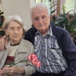 Starejši par sedi skupaj, gospa drži nagelj, gospod jo objema čez rame; oba se prijazno gledata v kamero.