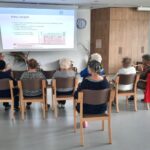 Skupina starejših oseb sedi na stolih v sejni sobi in posluša predavanje. Na platnu je prikazana projekcija z naslovom »Komu verjeti?«