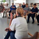 Predavateljica sedi obrnjena proti skupini starejših oseb, ki pozorno poslušajo. Ljudje sedijo v krogu, predavateljica govori in krepi svoje besede z gesto roke.