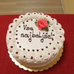 Okrogla torta z napisom »Vse najboljše!«, okrašena s smetano, rdečimi pikami in rožnato cvetlico na vrhu.