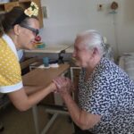 Klovnesa Rdeči noski v rumeni obleki drži za roke gospo v postelji, obe se nasmehneta.