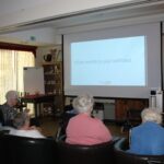 Skupina stanovalcev doma za starejše sedi v dvorani in gleda projekcijo na platnu, na katerem piše vprašanje v angleščini: "What month is your birthday".