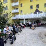 Starejši stanovalci doma upokojencev sedijo zunaj pred rumeno stavbo in jedo sladoled, nekateri uporabljajo hodulje ali invalidske vozičke.