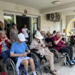Skupina starejših oseb v invalidskih vozičkih in na stolih pred domom upokojencev uživa sladoled.