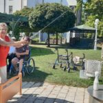 Dva stanovalca skupaj držita kroglo in merita proti kegljem na prostem, obkrožena z drugimi, ki jih opazujejo.