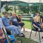 Skupina stanovalcev sedi na klopcah pod senčniki na vrtu doma in spremlja športne igre.