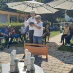 Stanovalec in stanovalka skupaj držita kroglo pri igri kegljanja na prostem, medtem ko ostali udeleženci v ozadju spremljajo dogajanje.