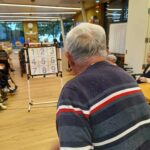 Starejši moški stoji pred tablo z devetimi številkami, pripravljen na met žogice, medtem ko drugi stanovalci opazujejo igro.