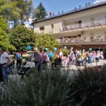 Skupina stanovalcev doma za starejše v invalidskih vozičkih z modrimi baloni, ob spremstvu svojcev in zaposlenih, na dvorišču pred domom.