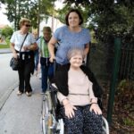 Stanovalka doma v invalidskem vozičku med sprehodom, ki jo spremlja ženska. Za njima hodijo drugi udeleženci sprehoda, med njimi tudi moški s palico.