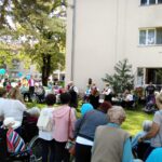 Večja skupina stanovalcev doma, svojcev in obiskovalcev se je zbrala na vrtu doma, kjer nastopa skupina žensk z glasbili. Nekateri stanovalci imajo v rokah modre balone.