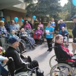 Stanovalci doma v vozičkih in na stolih ter svojci poslušajo nagovor zaposlene ženske, ki stoji z listom papirja pred smreko. Večina udeležencev drži modre balone.