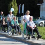 Skupina stanovalcev v invalidskih vozičkih z spremljevalci se sprehaja po cesti, v ozadju so avtomobili in stavba. Nekateri nosijo modre balone.