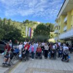 Velika skupina stanovalcev doma, zaposlenih in prostovoljcev stoji pred domom, nekateri v invalidskih vozičkih. Ob strani plapola slovenska zastava. Dan je sončen z modrim nebom.