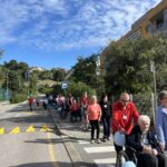 Udeleženci pohoda v vrsti na pločniku pred domom. Nekateri sedijo v vozičkih, drugi hodijo ob njih. V ospredju starejši gospod v rdečem puloverju potiska voziček gospe z modrim balonom.