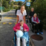 Ženska stoji za starejšo gospo v vozičku z modrim balonom v naročju. V ozadju so drugi stanovalci in prostovoljci ob cesti pri avtobusni postaji.