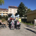 Skupina stanovalcev doma starejših v invalidskih vozičkih, ob njih zaposleni in spremljevalci. Vsi imajo modre balone, na dvorišču pred stavbo doma.