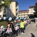 Dolga vrsta stanovalcev v invalidskih vozičkih z modrimi baloni, ki jih spremljajo zaposleni v rumenih brezrokavnikih, pred vhodom doma starejših na Ptuju.