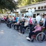 Skupina stanovalcev v invalidskih vozičkih in njihovih spremljevalcev, ki držijo modre balone, stoji na pločniku ob domu starejših.