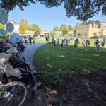 Stanovalci v invalidskih vozičkih z modrimi baloni opazujejo nastop otrok na travniku, pred vrtcem ob domu starejših.