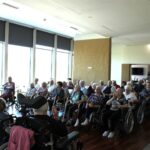 Skupina stanovalcev doma za starejše sedi v dvorani in posluša predavanje fizioterapevta.