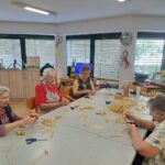 Skupina starejših žensk sedi za mizo in ustvarja izdelke iz ličja, okoli njih so škarje, slama in drugi materiali.