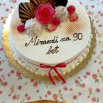 Okrugla torta z napisom »Mirandi za 90 let«, okrašena s sadjem in rožami, stoji na cvetličnem prtu.