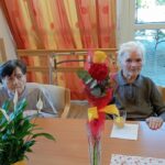 Stanovalci sedijo za mizo z rožami in sokom, ob praznovanju rojstnega dne v domu.