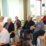 Skupina oseb sedi v dvorani doma, pozorno posluša in se nasmejano pogovarja.